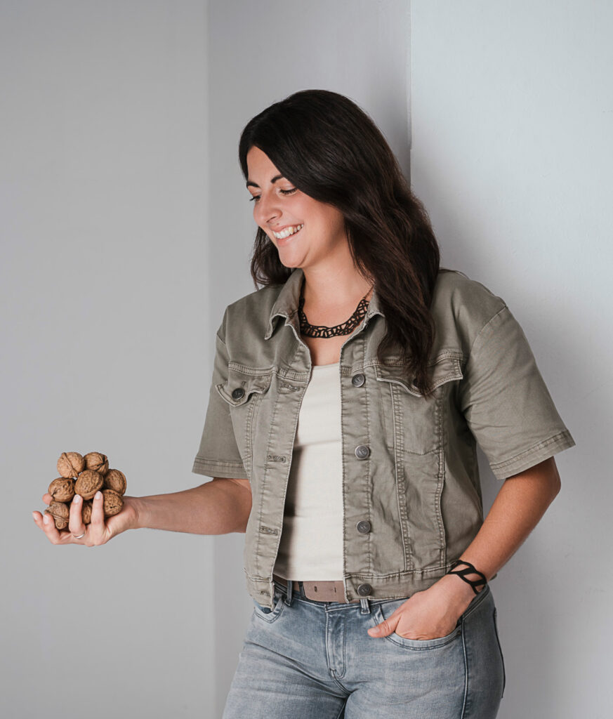 Nicole Klinglmair in einer khakifarbenen Jacke und Jeans lächelt, während sie eine Handvoll Walnüsse betrachtet.