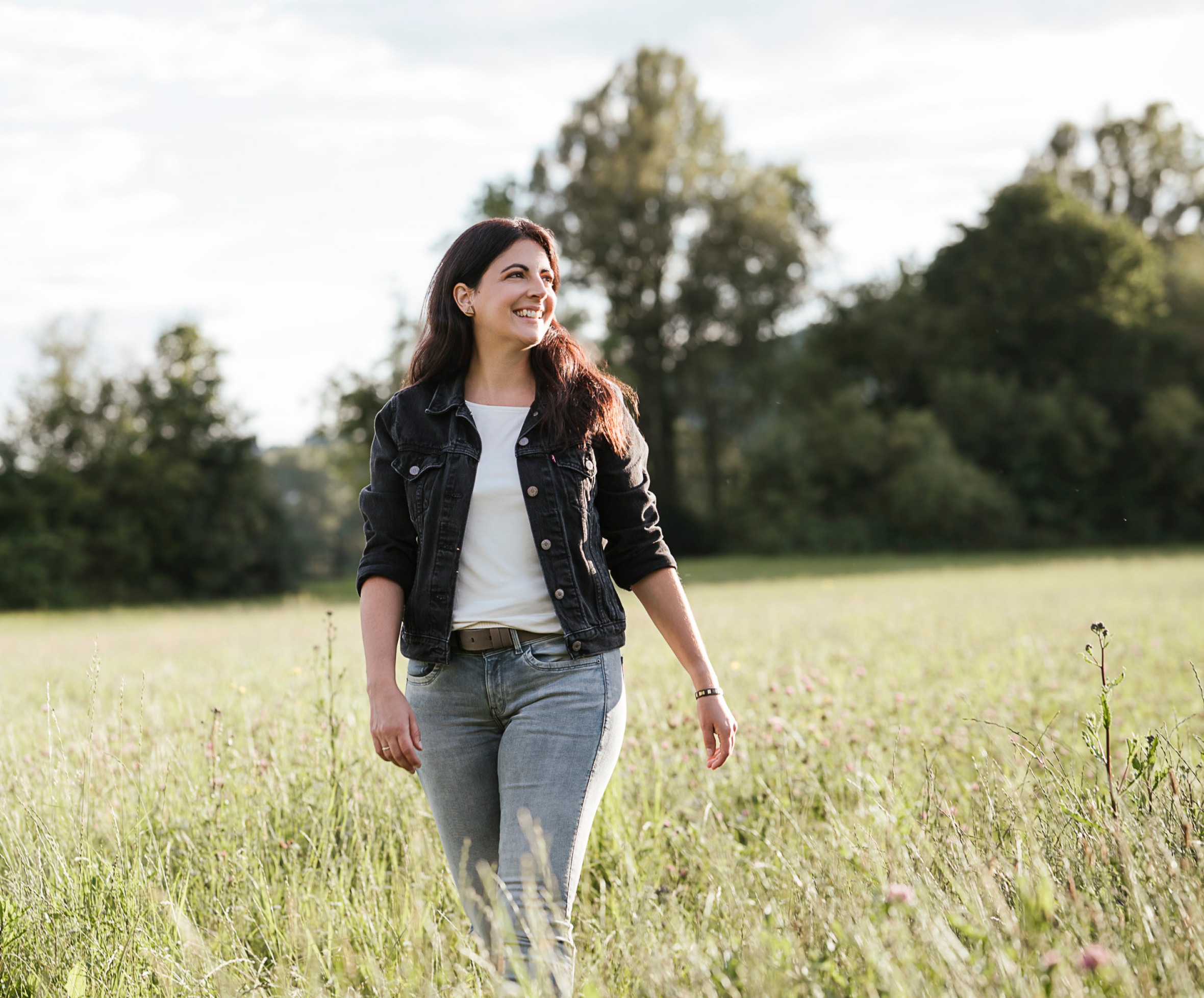 Nicole Klinglmair steht im Sonnenlicht auf einer grünen Wiese und blickt in die Ferne. Sie trägt eine schwarze Jacke und Jeans.
